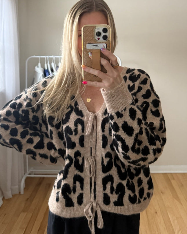 Person wearing a leopard print sweater taking a mirror selfie in a room with a window and wooden floor.
