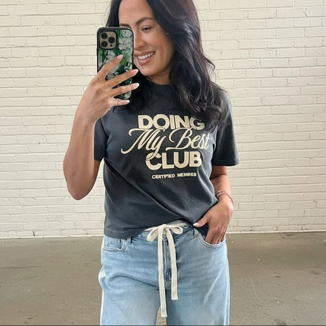 Woman taking a mirror selfie wearing a 'Doing My Beauty Club' t-shirt in a store setting.