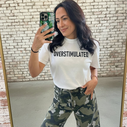 Person wearing a white t-shirt with 'OVERSTIMULATED' text and camouflage pants, taking a mirror selfie.