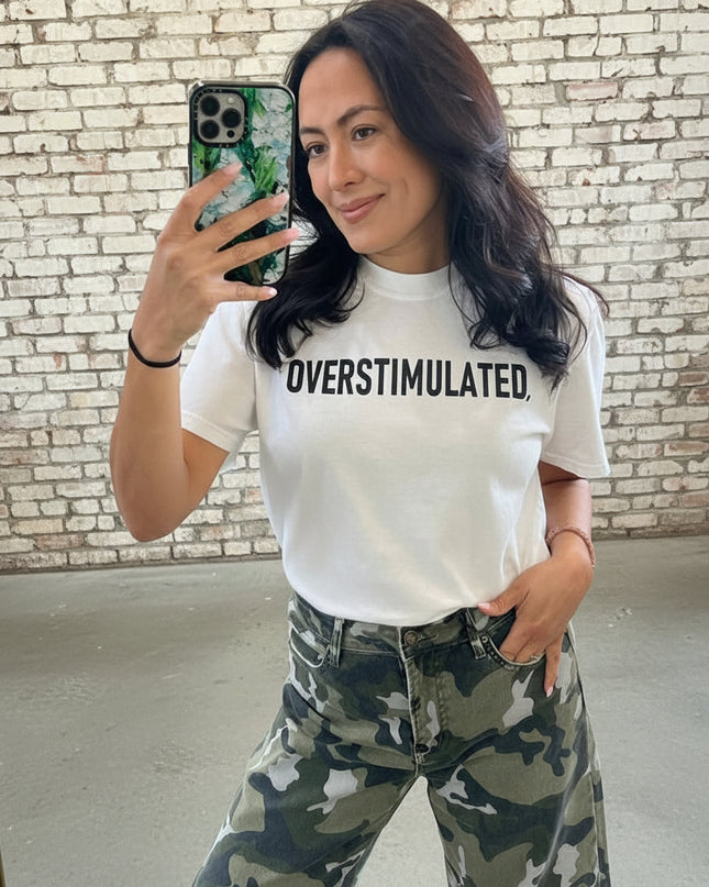 Person wearing a white t-shirt with 'OVERSTIMULATED' text and camouflage pants, taking a mirror selfie.